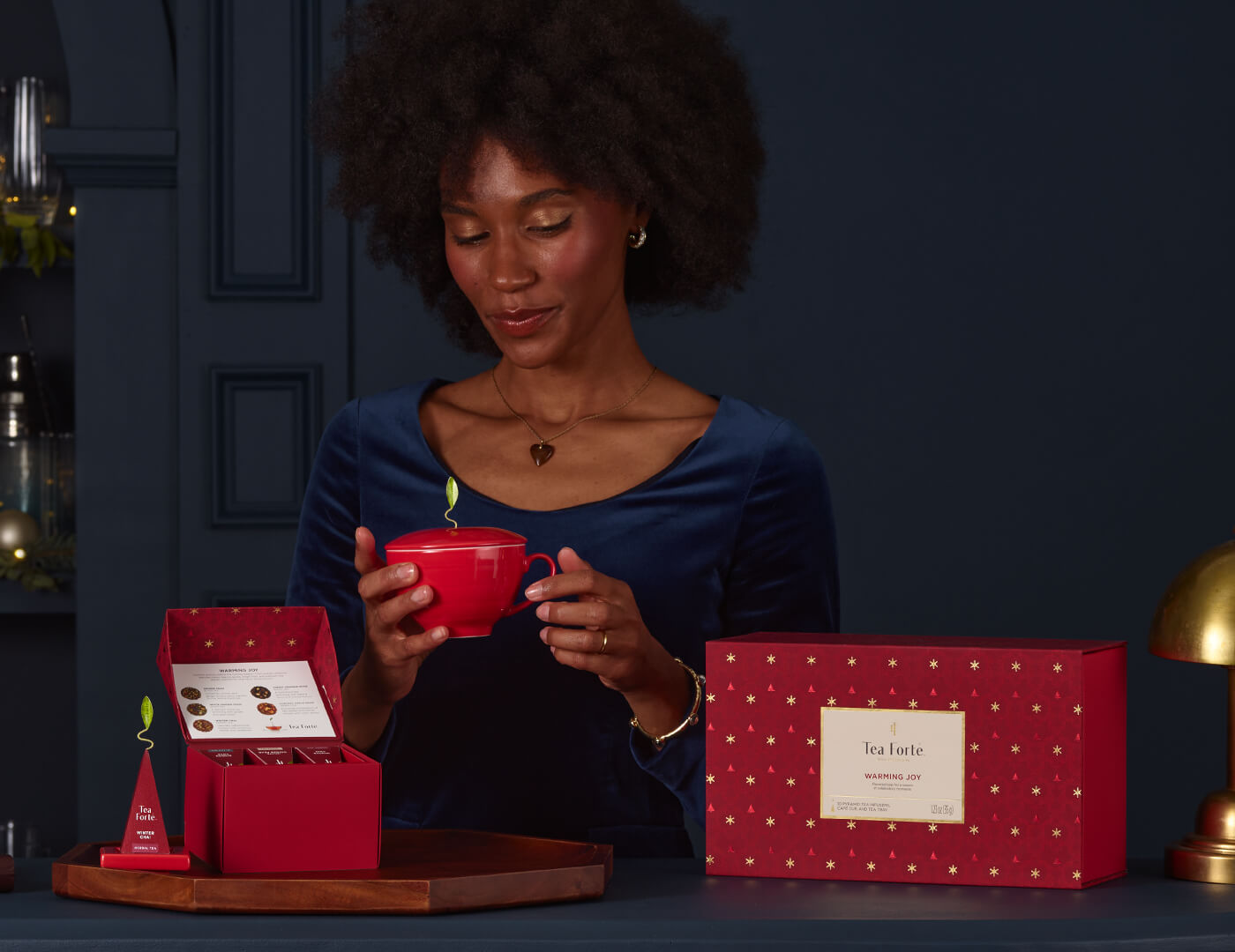 Woman holding a red mug of the Warming Joy Gift Set, surrounded by tea packaging on a dark blue background